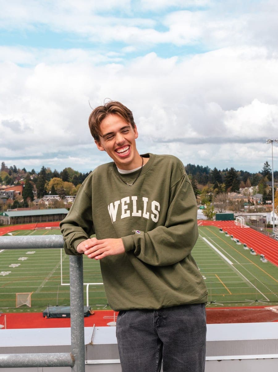 Student laughing in an olive Wells crewneck above the school’s track and field