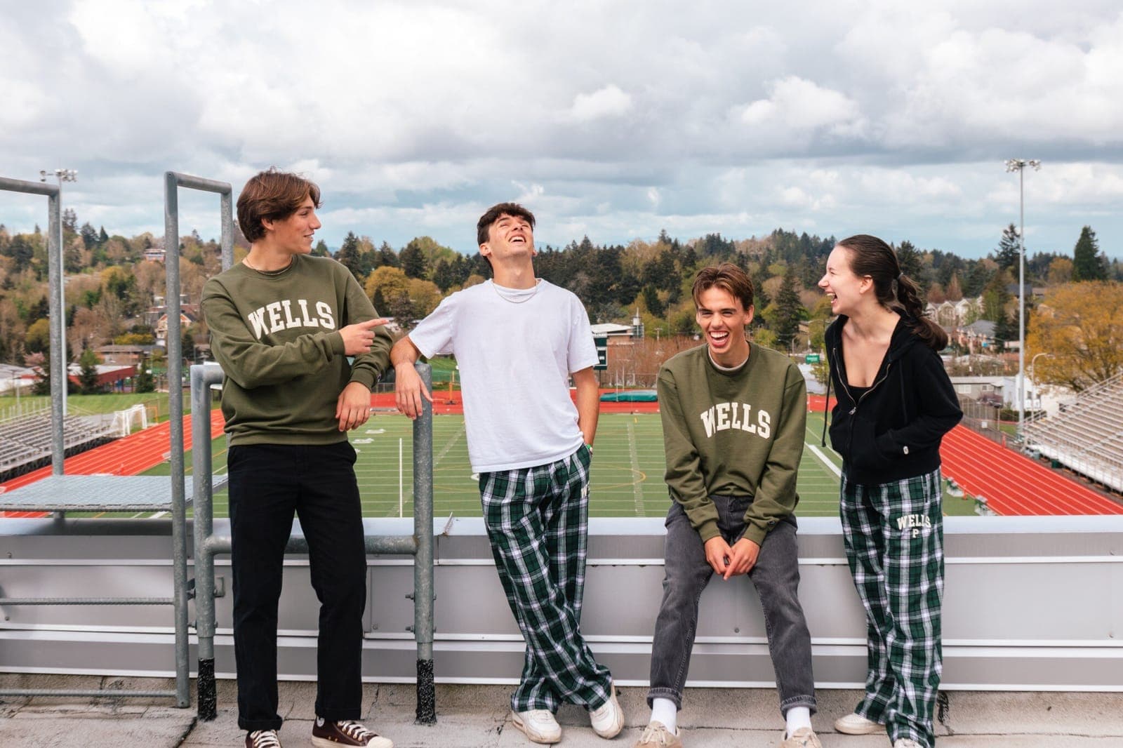 Four students laughing on a stadium rooftop wearing the Wells merchandise line, with the school’s track and field below